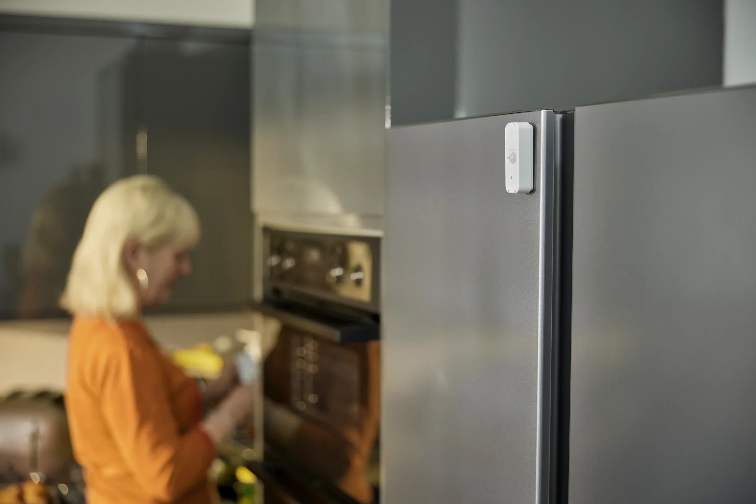 Older woman in the kitchen next to Canary Care sensor on the fridge, showing how smart home monitoring tech monitors activity.