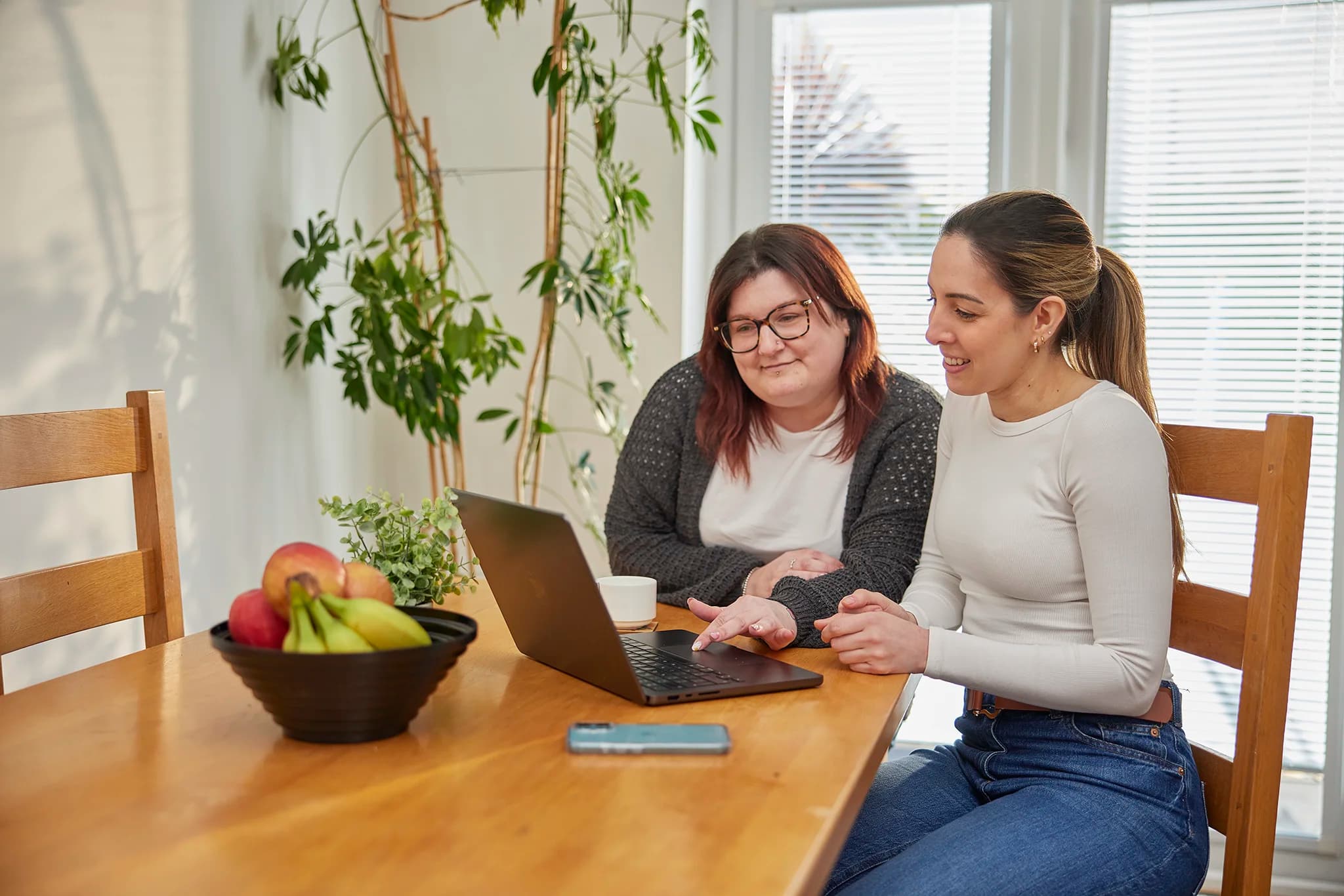 Two women smiling and using a laptop at a kitchen table, symbolising Canary Care's user-friendly smart home monitoring tech.